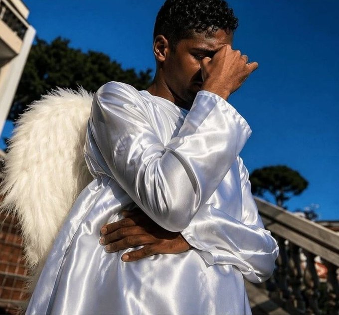 A man wearing a white satin shirt with fluffy white angel wings stands outdoors with his hand to his face, set against a bright blue sky.