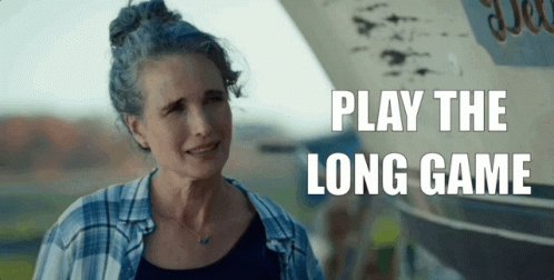 The image features an older woman with gray hair wearing a blue shirt, standing next to a boat or ship. The text overlaying the photo reads "Play the long game", which seems to be a part of a larger advertisement or promotional material for a project that is focused on building infrastructure rather than chasing narratives.