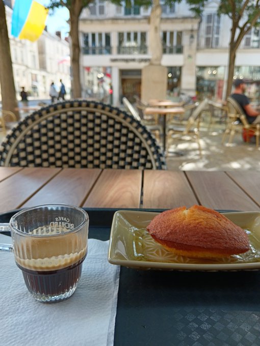 An espresso macchiato in a clear glass cup and a golden-brown madeleine pastry on a square green plate sit on a wooden table at an outdoor cafe, with a checkered chair and city square visible behind.