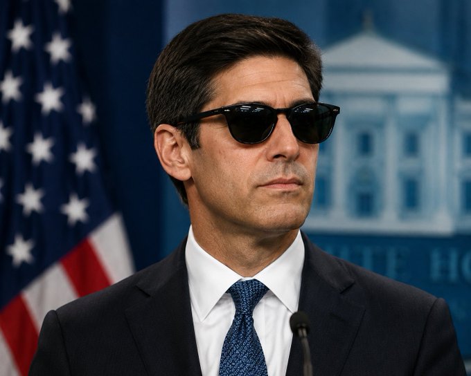 A man in sunglasses and a suit stands at a White House press briefing podium with an American flag behind him, his expression stoic. The image appears to comment on the post's discussion of inflation data, with the sunglasses symbolically "shading" the viewer's view of the core CPI beat, suggesting an attempt to downplay the unwelcome economic news for Bitcoin ahead of the FOMC meeting.