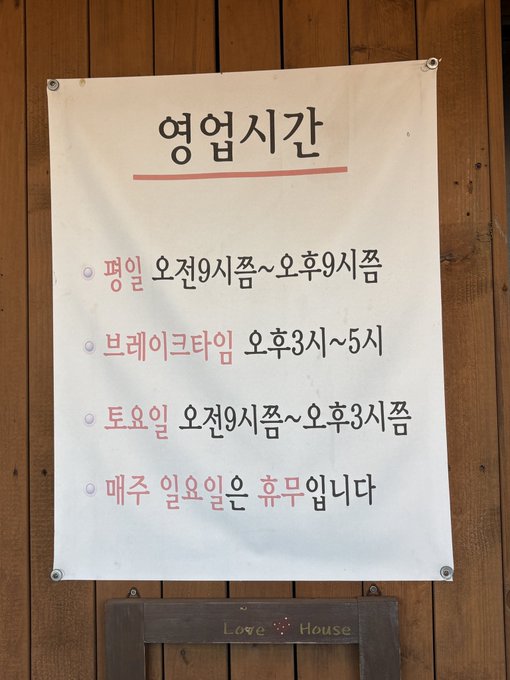 A sign in Korean titled '영업시간' (Business Hours) mounted on a wooden wall, listing hours for weekdays (9 AM-9 PM), break time (3-5 PM), Saturdays (9 AM-3 PM), and noting closed on Sundays and public holidays.