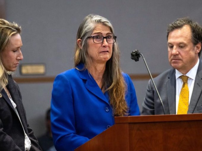 This image does not contain any visible text, numbers, or names that I can identify. I can only describe what I observe visually.

The image shows a woman in a blue blazer and glasses standing at a podium with a microphone, flanked by two other people. To her left is a person with long hair in a dark jacket, and to her right is a man wearing a gray suit and yellow tie. The woman at the center appears to have a serious or emotional expression, and the setting suggests a courtroom or formal proceeding.