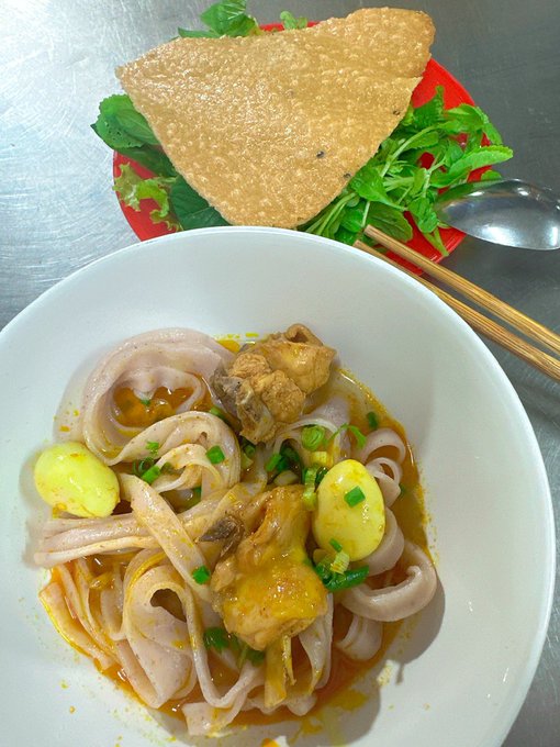 A bowl of noodle soup with chicken and quail eggs served alongside a side plate of fresh greens and a crispy fried wonton wrapper.