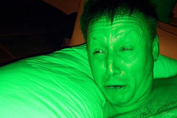 A close-up photo of a man's face illuminated by green light, lying down on a pillow with a 'Grok' watermark in the corner.