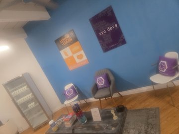 A room decorated for xDay Devs, featuring a small refrigerator with xDay branding, purple xDay mugs on shelves, and posters on the blue walls.