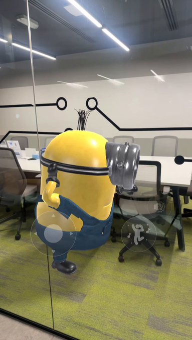 A Kevin the Minion mascot stands in an office conference room featuring a glass wall decorated with a circuit-board pattern and a meeting table with office chairs. The image likely hints at playful or quirky updates from the company, with Kevin appearing to peer curiously into the workspace, suggesting something new is "brewing" behind the scenes.