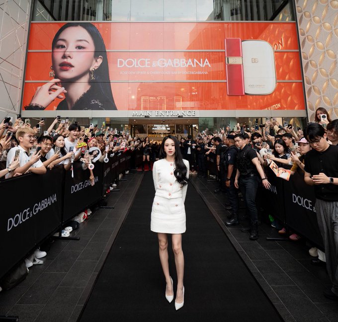 A photograph of an Asian woman in a white outfit walking on a black carpet at a Dolce & Gabbana event, flanked by crowds of fans behind barriers, with a large red Dolce & Gabbana cosmetics advertisement displayed on the building behind her.