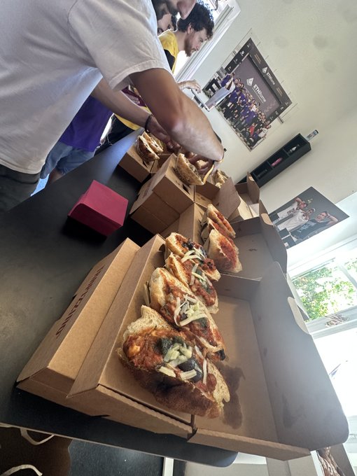 Several open pizza boxes containing slices topped with cheese, herbs, and vegetables are arranged on a dark table, with a person in a white shirt reaching across the scene.