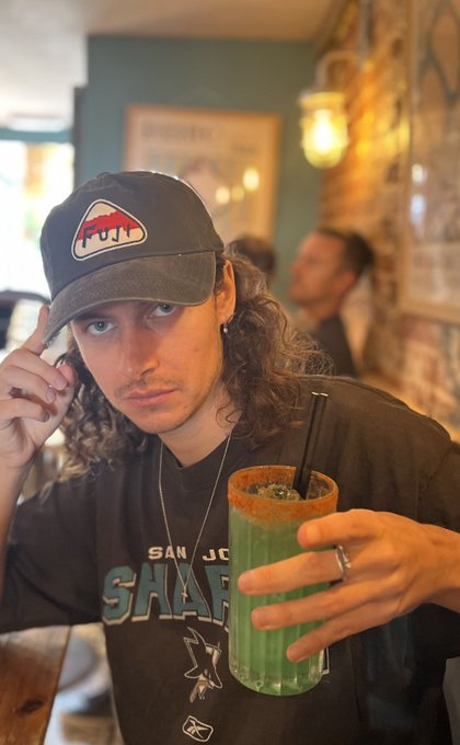 A young man wearing a black San Jose Sharks cap and t-shirt, holding up a green Michelada with a chili-salt rim.