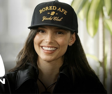 A woman smiles while wearing a black baseball cap embroidered with "BORED APE YACHT CLUB" in gold lettering, fitting the post's playful "this is me, this is my ape" comparison between herself and a Bored Ape NFT holder.