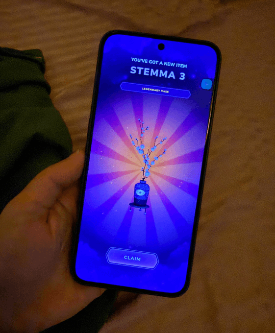 A hand holding a smartphone displaying a "YOU'VE GOT A NEW ITEM" screen for a "STEMMA 3" item with a "LEGENDARY RARE" label and a "CLAIM" button, set against a purple background.