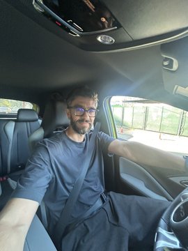 A selfie of a man with glasses and a beard sitting in the driver's seat of a car.