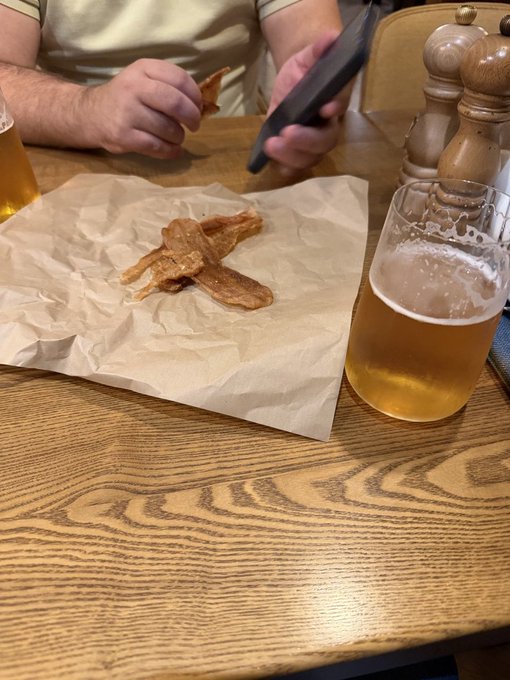 A person at a wooden restaurant table holds a phone in one hand and a piece of fried food (possibly fish skin or chicharron) in the other, with the remaining food on brown parchment paper next to a glass of beer.