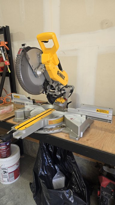 A yellow DeWalt miter saw sits on a wooden workbench in a garage or workshop setting, with construction materials and buckets nearby.