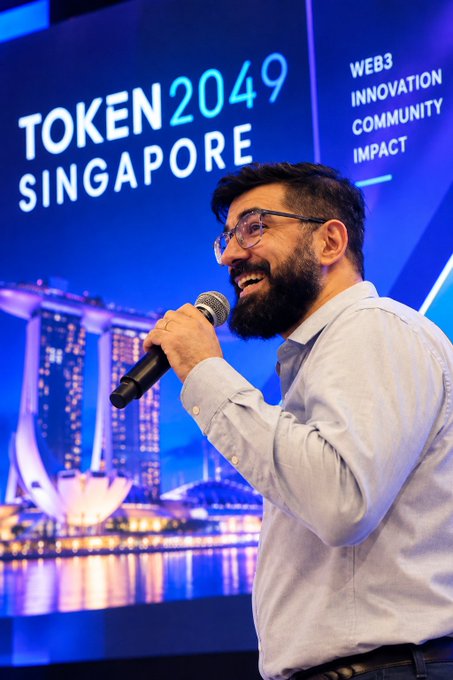 A man wearing glasses speaks into a handheld microphone on a stage, smiling in front of a blue screen displaying “TOKEN2049 SINGAPORE,” along with the words “WEB3,” “INNOVATION,” “COMMUNITY,” and “IMPACT.” The scene visually matches the post’s imagined scenario of Nicolas Kokkalis appearing at TOKEN2049 Singapore 2026 to discuss Pi Network and Web3.