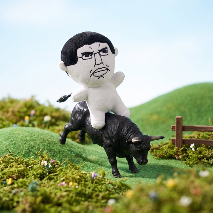 A whimsical figurine of a plush character with black hair and glasses riding a black bull through a miniature green landscape with hills, flowers, and a small wooden fence.