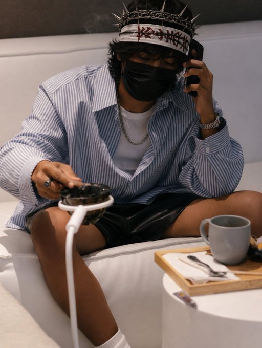A young man wearing a black face mask and a spiked headband sits on a white couch, holding a phone to his ear while operating a hookah with his other hand.