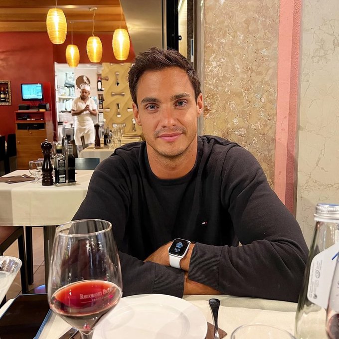 A man in a black Tommy Hilfiger sweater sits at a restaurant table with a glass of red wine, wearing a smartwatch, in what appears to be an Italian restaurant with a chef visible in the background.
