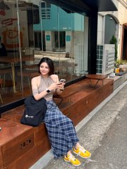 A young woman with dark hair stands outdoors on a sidewalk, wearing a sheer top, a blue plaid skirt, and yellow sneakers, while holding a phone and carrying a black tote bag.