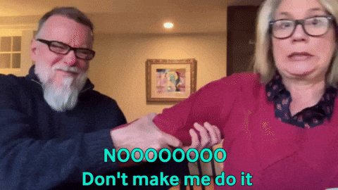 The image features a man with a beard wearing glasses, and a woman with blonde hair wearing glasses. They are both smiling at the camera while holding hands. Below them, there's text that reads "NOZZZOO! DON'T MAKE ME DO IT". The background of the image is blurred, but it appears to be a room or a living space.