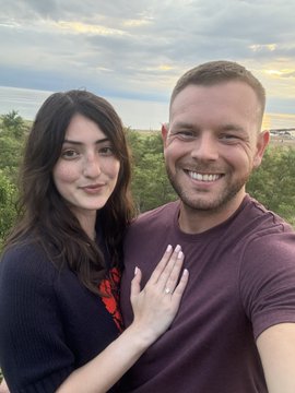 A selfie of a smiling couple at sunset, with the woman showing off an engagement ring on her finger.