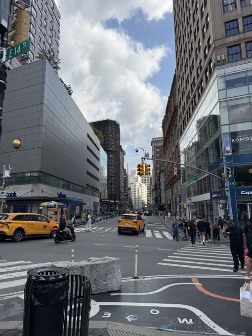 A street view of New York City showing yellow taxis, pedestrians, and a visible 'E 14 St' street sign under a cloudy sky.