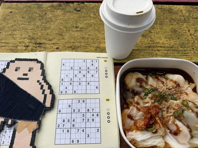 A top-down photo of a coffee cup, an open Sudoku puzzle book, a pixel-art coaster, and a bowl of rice noodle rolls topped with chili and scallions.