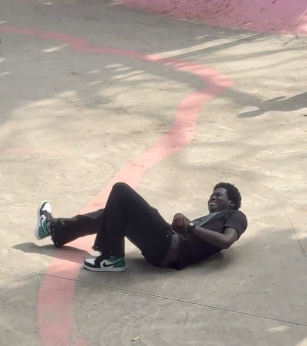 A young man lies on the ground at a skatepark, wearing black jeans, a dark t-shirt, and green and white sneakers, while laughing with his head tilted back. The casual, relaxed pose fits the upbeat "good morning" vibe of the vlog promo, which teases discussion of $RIVER and grassmax with Spenda later in the day.