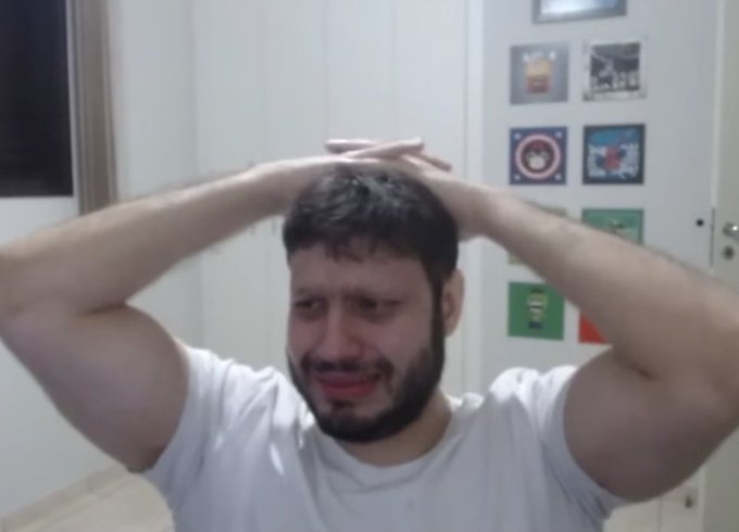 A man with dark hair and a beard, wearing a white t-shirt, holds his hands on his head with a distressed expression, reacting to accidentally exposing his crypto wallet seed phrase on a live stream and losing $60,000 in savings.