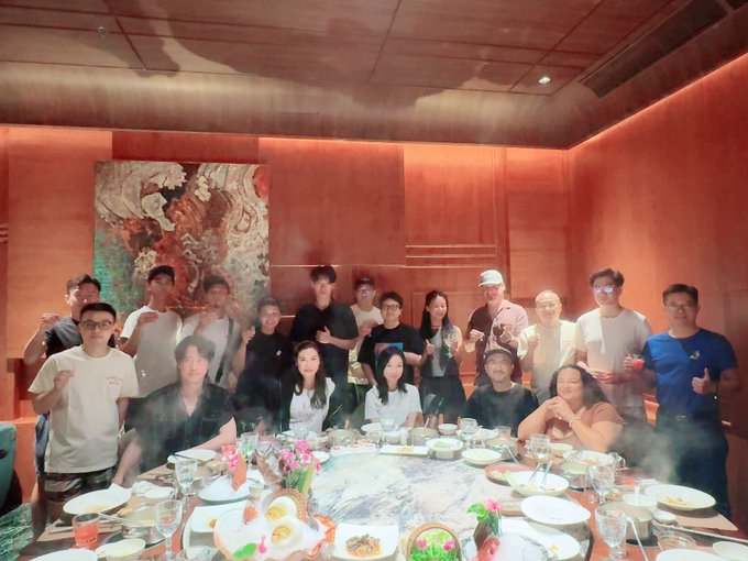 A group photo of people gathered around a large round dining table in a warmly lit private dining room with traditional artwork on the walls.