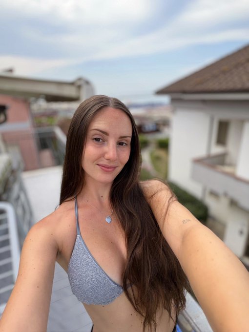 A woman with long brown hair wearing a gray bikini top takes a selfie on a balcony with a residential neighborhood in the background, capturing a relaxed morning vibe to match her cheerful "Good morning fam! Happy Thursday" greeting.