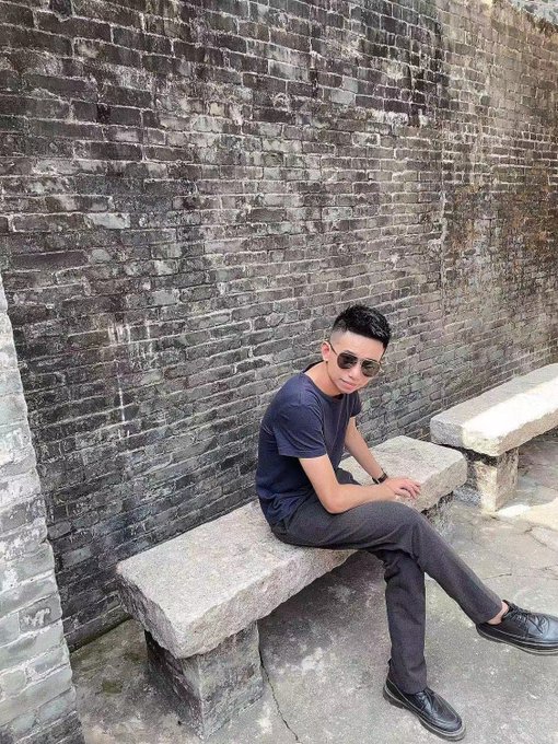 The image features a man sitting on a stone bench, wearing sunglasses and a blue shirt. The photo has been edited with a filter that gives it a vintage appearance, making the man look older than he actually is. The background of the image consists of a brick wall, which adds to the overall aesthetic of the scene.
