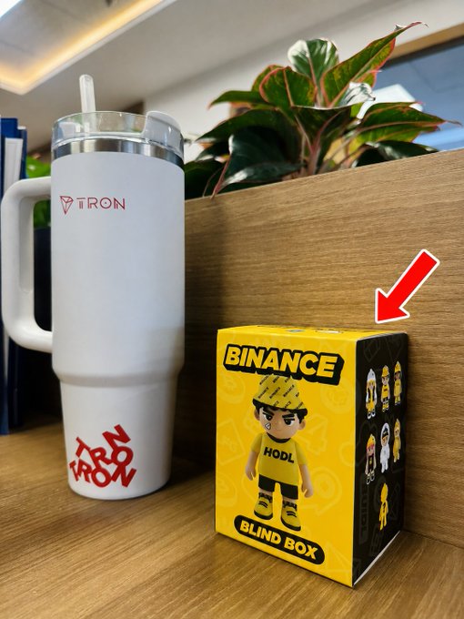 A white travel tumbler with 'TRON' and 'WE ARE HODL' logos sits next to a yellow 'BINANCE BLIND BOX' box with a 'HODL' character, indicated by a red arrow.
