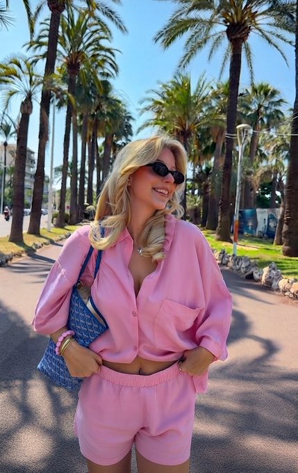 A blonde woman wearing sunglasses and a pink shirt outfit, standing on a palm-tree-lined promenade.