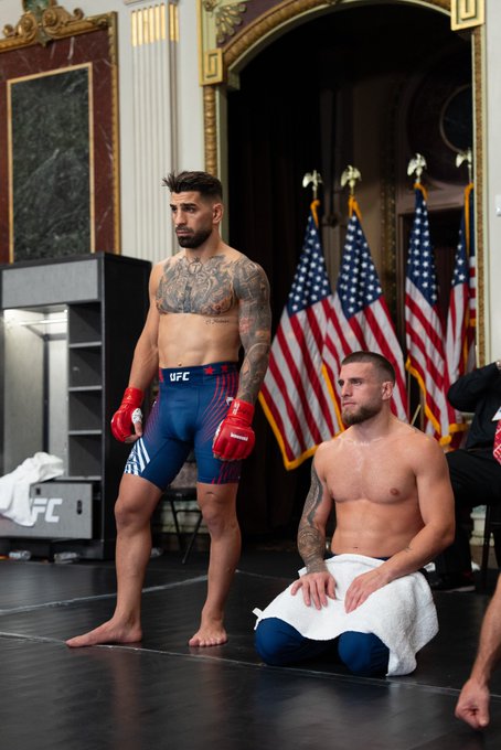 The image shows two men standing next to each other, one of them wearing boxing gloves while the other has a towel wrapped around his waist. The man with the towel appears to be kneeling down, possibly resting or preparing for their training session. The background features an American flag and a cabinet, suggesting that they are in a room related to the UFC or a similar organization. The image also contains text elements such as "UFC White House", "@PPlusDeportes", "#UFCWhiteHouse", and "@cryptocom".