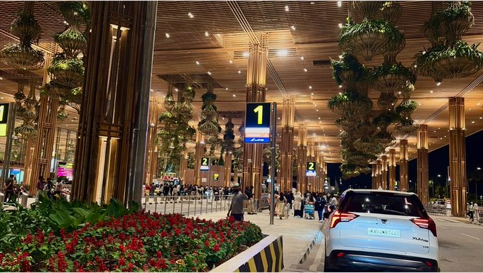 A nighttime scene at an airport arrivals area featuring a covered walkway decorated with hanging plants and red flower beds, with taxi stand signs displaying numbers 1 and 2.