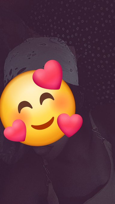 A selfie photo of a person whose face is obscured by a smiling emoji with hearts, set against a dark purple patterned background.