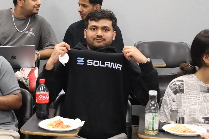 A young man smiles while proudly displaying a black "SOLANA" hoodie during what appears to be a casual gathering, with plates of pizza and soda bottles on the tables around him.
