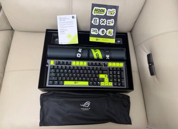 A boxed set of ROG (Republic of Gamers) keyboard and mouse accessories in black and neon green packaging, placed on a beige leather seat.