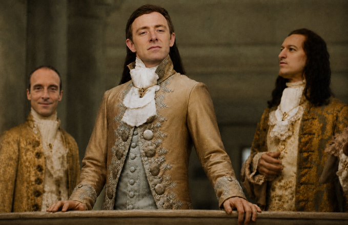 A man in an ornate tan and silver 18th-century period costume with a ruffled cravat stands at a balcony railing, flanked by two other men in similarly elaborate gold-toned aristocratic attire.
