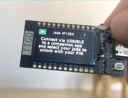 A hand holds a TTYGO Jade 4F10E0 development board displaying the prompt "Connect via USB/BLE to a companion app and select your Jade to unlock with your PIN." The image illustrates the DIY Jade hardware referenced in the post, showcasing the board's initial setup screen for connecting to the companion app as described in Blockstream's open-source guide.