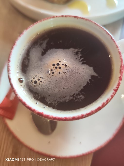 Close-up photo of a cup of dark coffee or tea with foam bubbles on the surface, served in a white cup with a red rim on a saucer, photographed with a Xiaomi 12T Pro by @SACRA97.