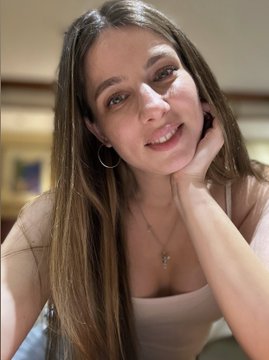 The image features a woman with long brown hair wearing a white tank top, smiling at the camera while resting her hand on her cheek. The background of the photo shows a wooden wall, suggesting that she might be indoors or in an artistic setting. The caption of the image reads "SOLD OUT Thank you so much @_bitcoiner for your support I’m incredibly happy that my art in your collection now Big hugs".