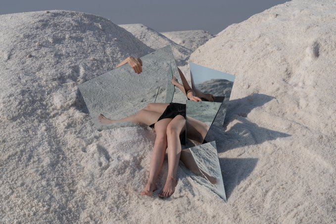 A surreal digital artwork showing a fragmented figure sitting against white, salt-like mounds, with mirrors reflecting different angles of the body and a bright sky above.