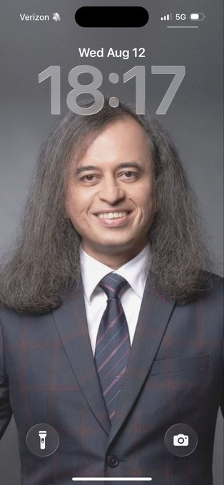An iPhone lockscreen display showing the date 'Wed Aug 12' and time '18:17' on a Verizon 5G signal, featuring a portrait of a smiling man with long dark hair wearing a suit and tie.