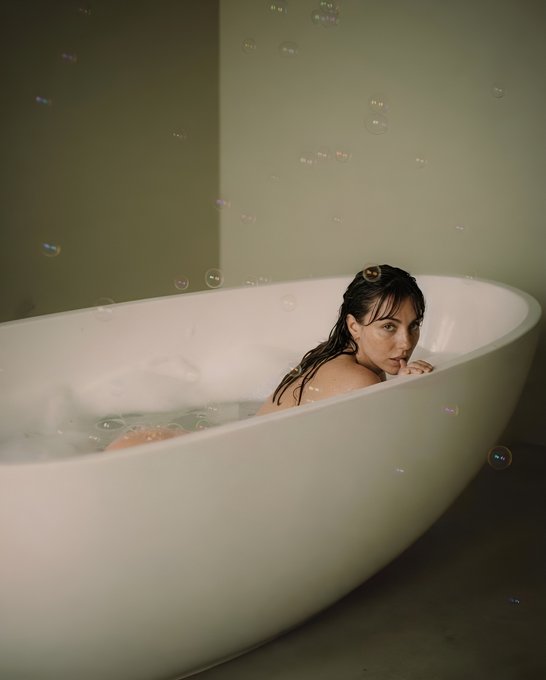 A moody portrait of a woman taking a bubble bath in a white freestanding tub, looking over her shoulder at the camera with bubbles floating in the air around her.