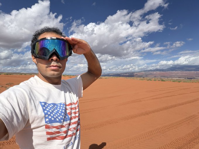 A selfie of a young man wearing ski goggles and an American flag t-shirt, standing in a desert landscape with red sand dunes under a blue cloudy sky.