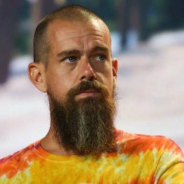 A close-up portrait of a bearded man with a shaved head wearing an orange and yellow tie-dye shirt.