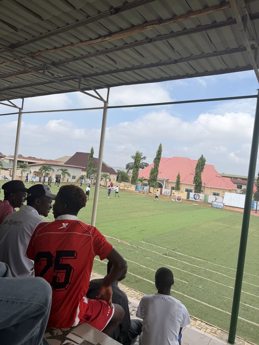 Fans wearing a jersey with "25" and another reading "Legally" watch from a covered stand as players compete on the field at the Ilorin Web3 Boys football match.