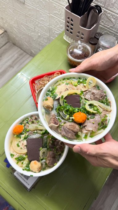 A close-up shot of someone holding two bowls of Vietnamese pho, a noodle soup with beef, bone marrow, and congealed blood, taken over a green wooden table with a chili condiment container.